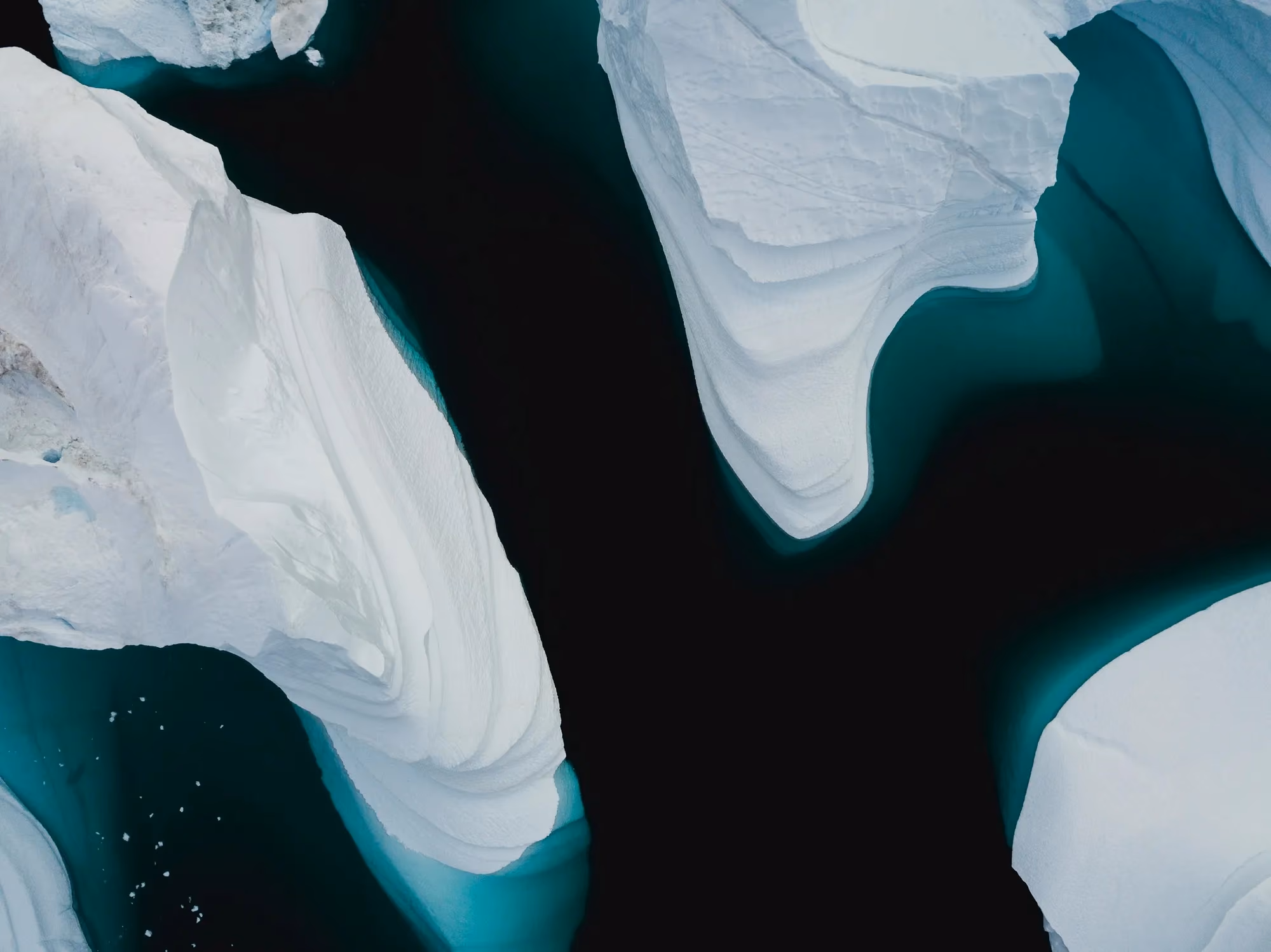An aerial view of arctic ice floes, white slabs of ice separated by channels of dark teal water, so a single frozen surface reads as broken into distinct pieces.