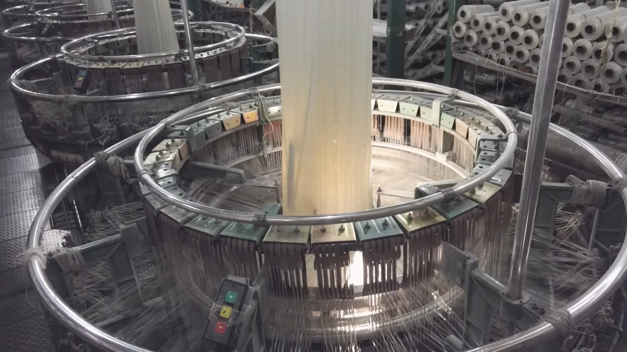 Two large circular looms on a factory floor, rings of shuttles weaving tubes of polypropylene fabric that rise toward the ceiling.